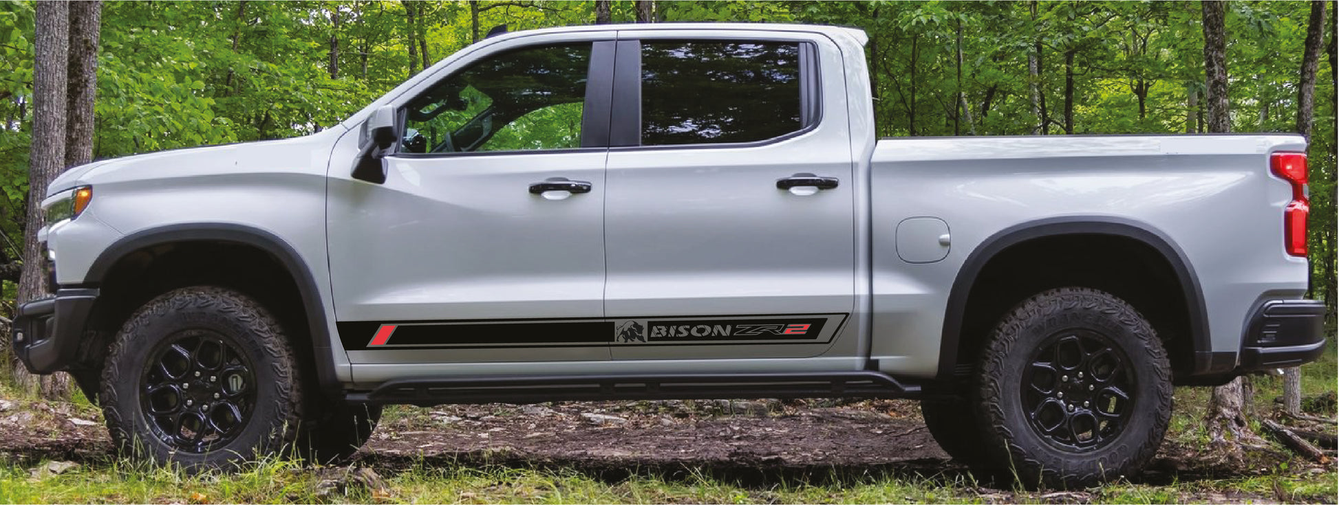 A white pickup truck shown in side profile, parked outdoors on a dirt path surrounded by green trees. The truck has black wheels, a black front bumper, and a black side decal with red accents.