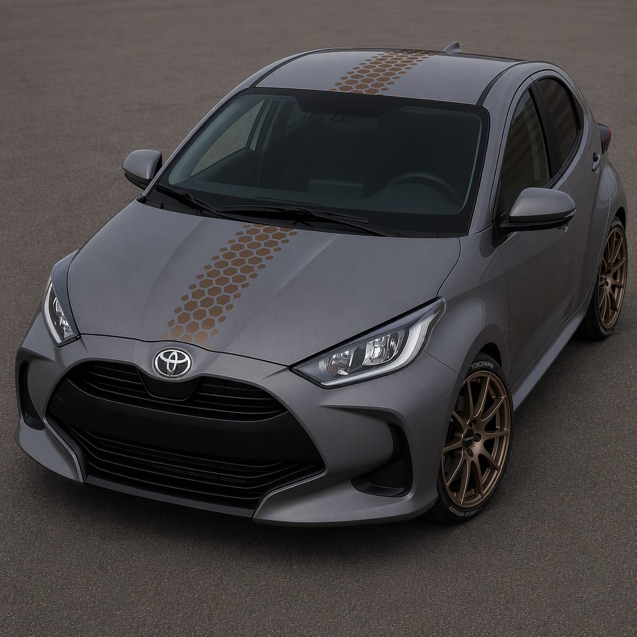 A gray Toyota hatchback car with a black honeycomb pattern decal running down the center of the hood and roof, parked on a gray asphalt surface.