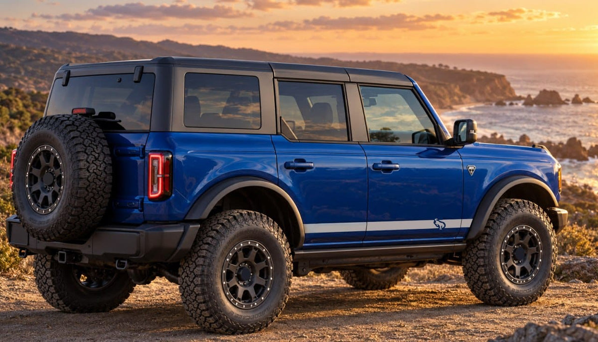 A blue Ford Bronco Sport SUV with black off-road tires and a spare tire mounted on the rear, parked on a rocky coastal path at sunset.