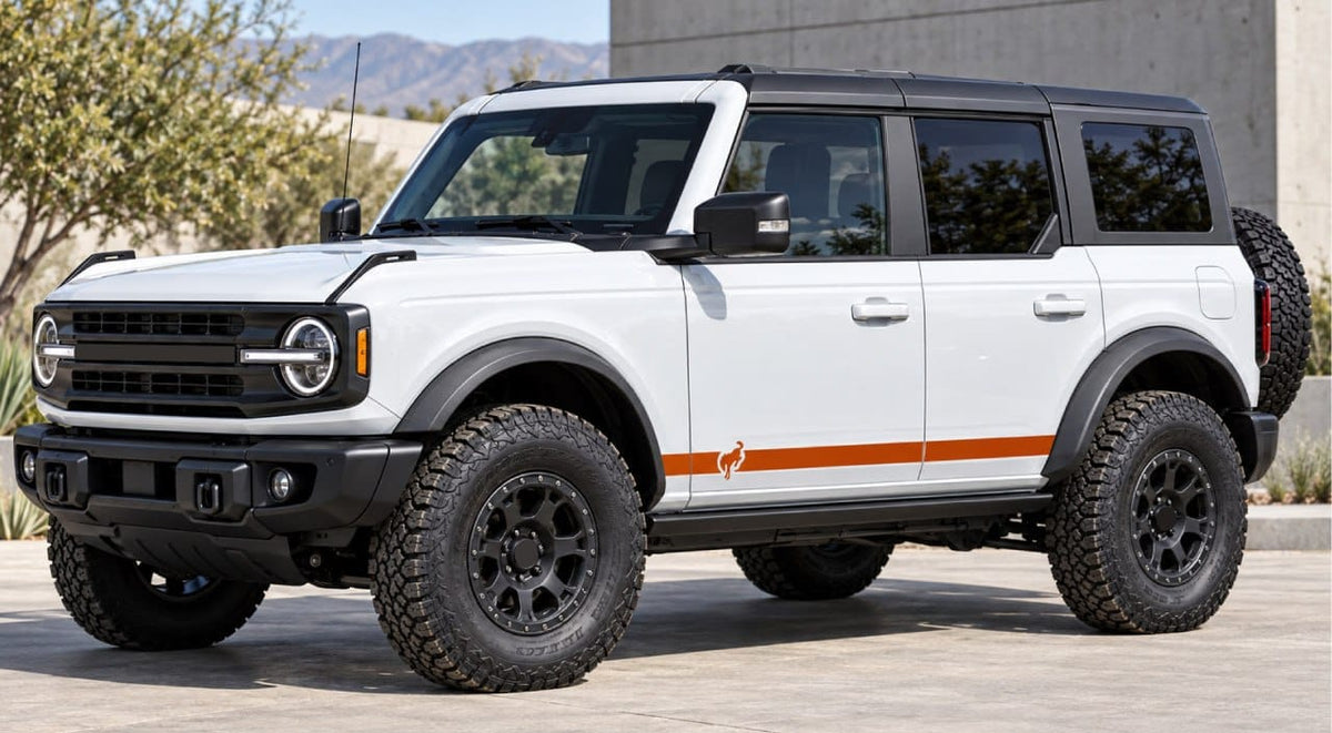 A white Ford Bronco with a black roof and black wheels, featuring a distinctive orange side stripe and a small orange fox logo, parked outdoors in front of a modern building.
