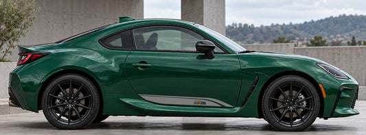 A dark green sports coupe car shown in a side profile view, parked outdoors near a concrete structure with hills in the background.