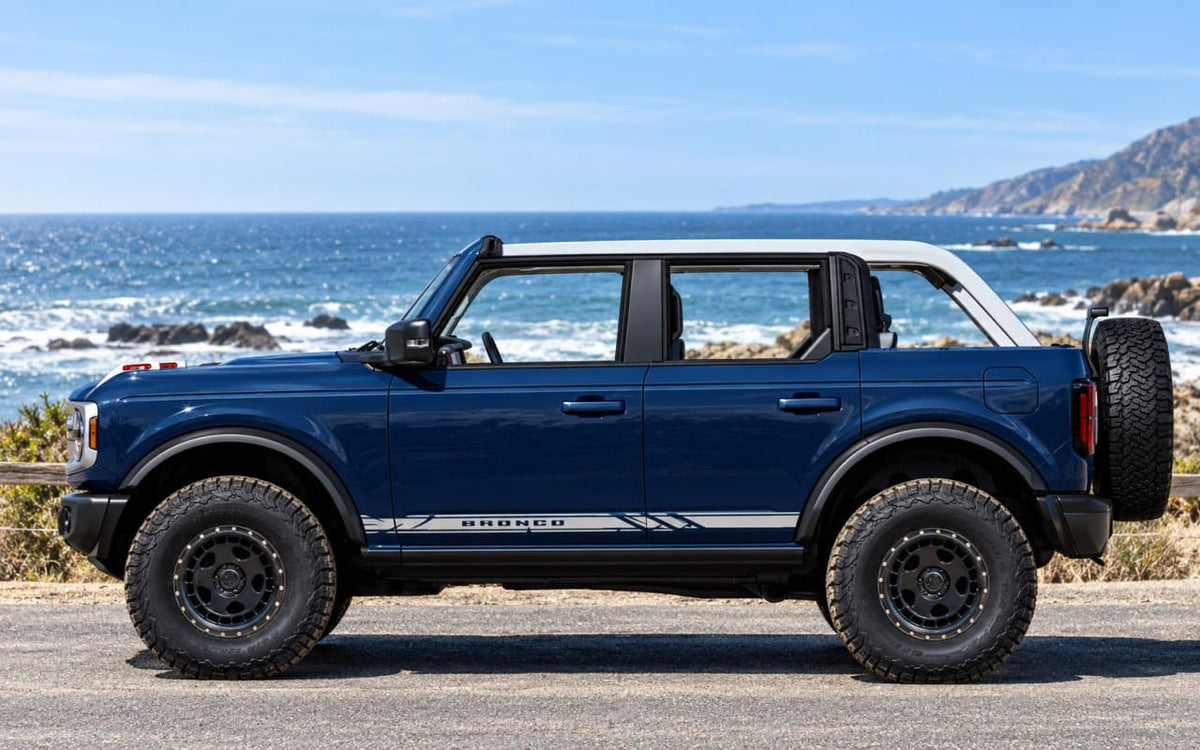 A blue Ford Bronco with a white roof and black off-road tires parked outdoors by the ocean, with rocky coastline and blue sky in the background.