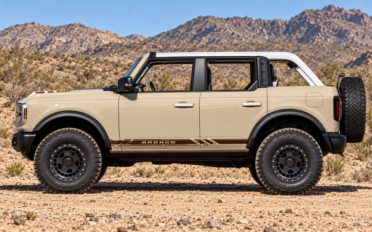 A tan Ford Bronco SUV with a white roof and black tires, parked in a desert landscape with rocky mountains in the background.