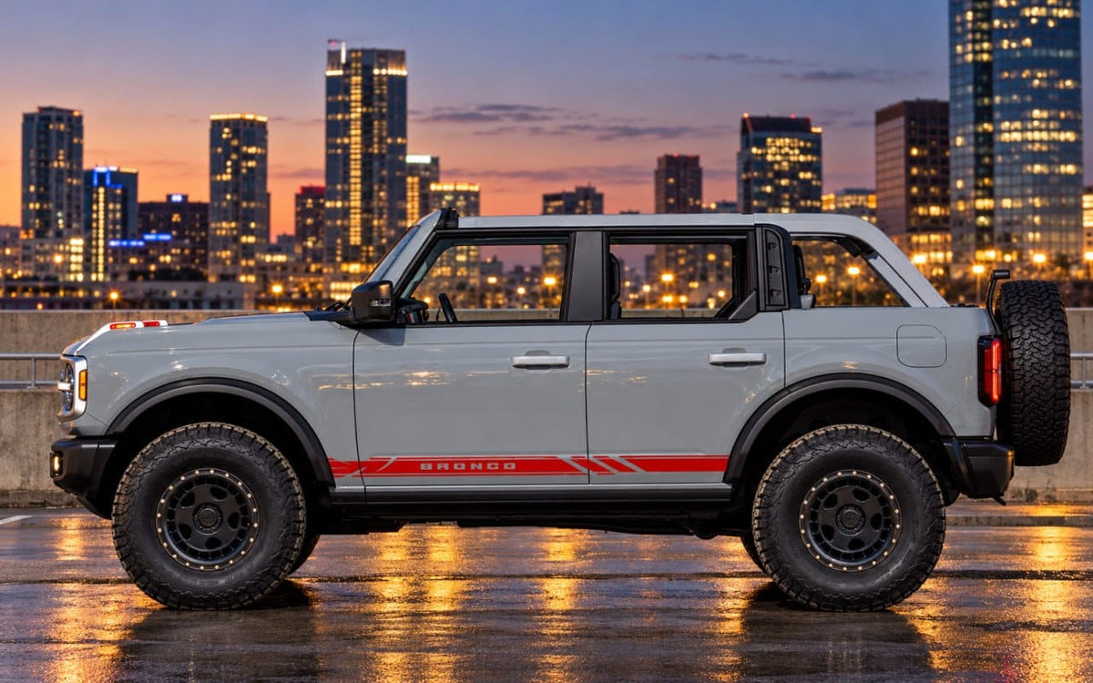 A light gray Ford Bronco SUV with a red side stripe and 'BRONCO' lettering, parked on a wet city street at dusk with a glowing skyline in the background.