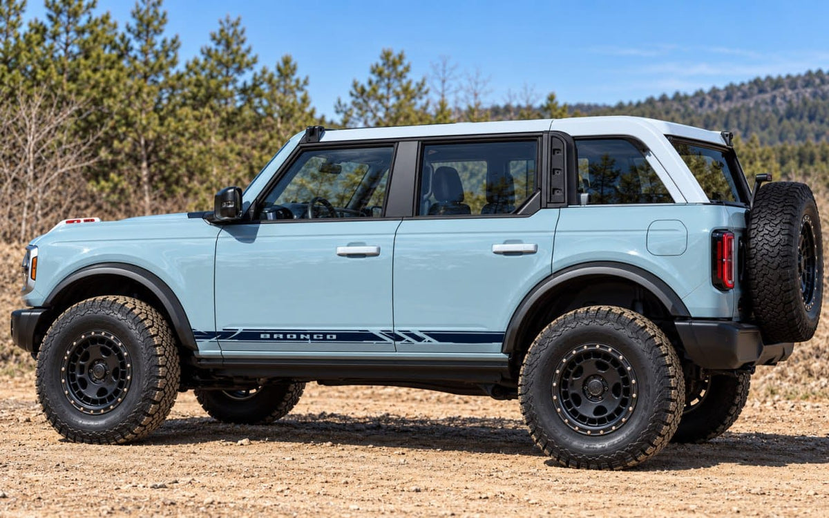 A light blue Ford Bronco with a white roof and black trim, parked outdoors on a dirt road with trees and hills in the background.